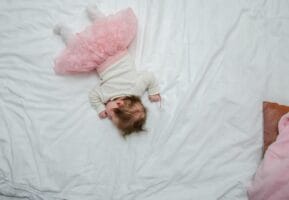 Η εξομολόγηση: Αγνή και τρυφερή εικόνα μωρού σε ροζ φόρεμα, ξαπλωμένο σε λευκό κρεβάτι."